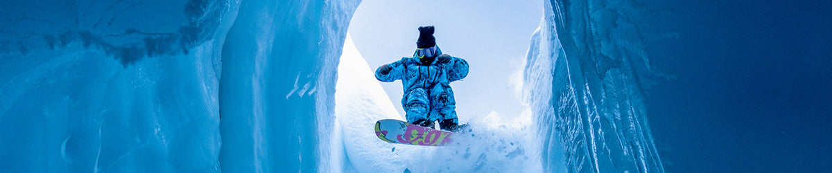 A snowboarder in a blue and white patterned snowsuit stands on a snowboard inside a bright ice cave, looking upward toward the opening.