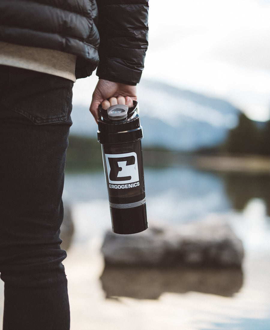A person outdoors by a lake holding a black water bottle with a white and black logo that reads EROGENICS.