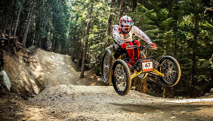 A mountain biker in a forest setting, riding a red and yellow trike with three wheels over a dirt trail. The rider is wearing a white and red helmet and protective gear.