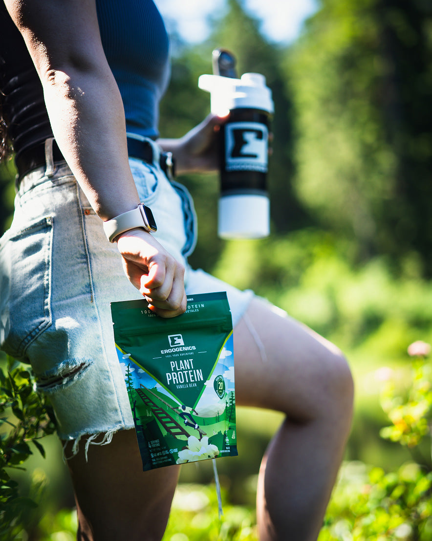 A person outdoors in a garden holding a green pouch of Plant Protein powder and a black shaker bottle with a white cap.