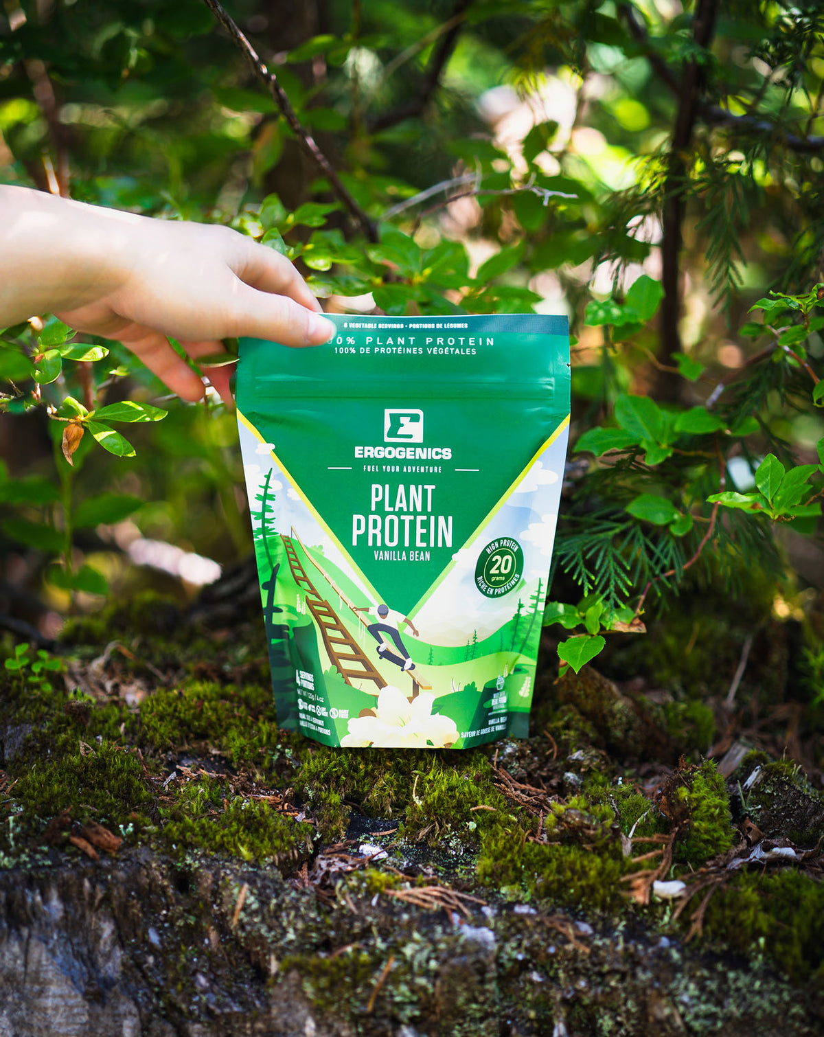 A hand holds a green Ergogenics Plant Protein powder pouch outdoors on a mossy log surrounded by green forest foliage.
