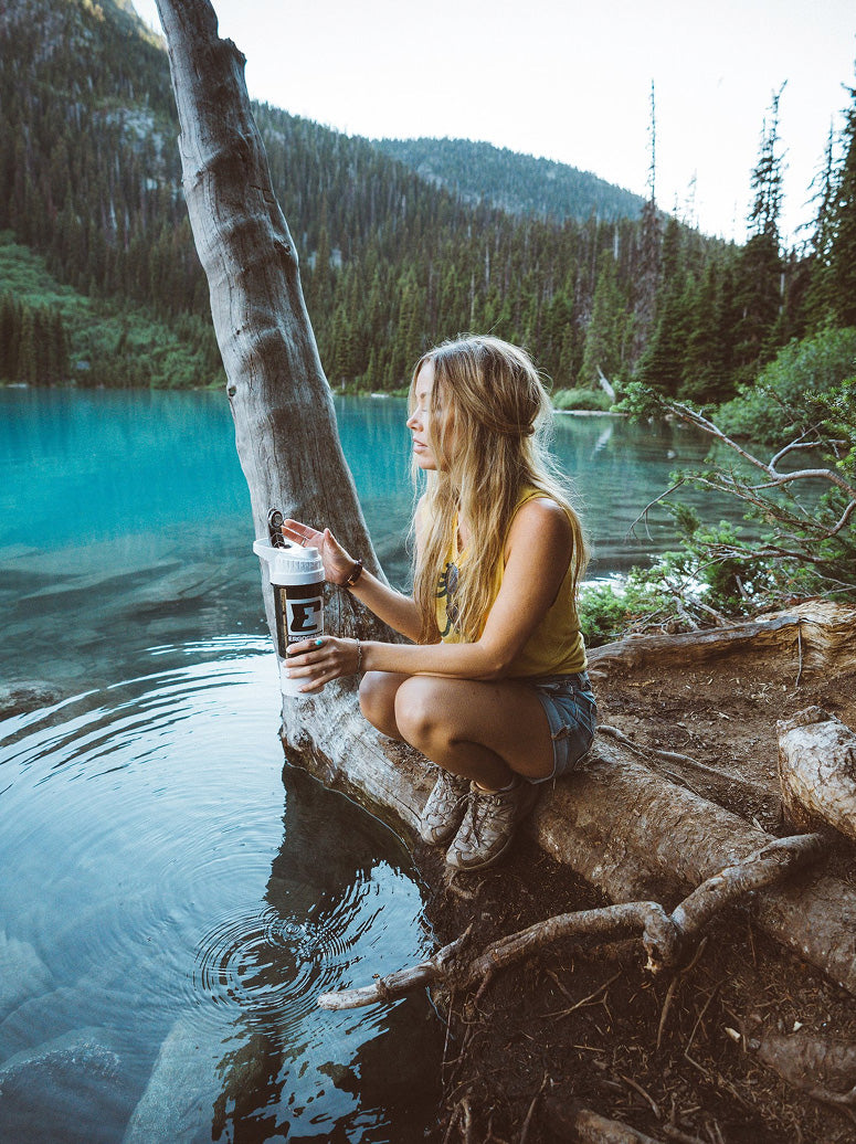 A woman sits on a log by a turquoise lake in a forested mountain setting, holding a coffee cup. She is wearing a yellow tank top and denim shorts.
