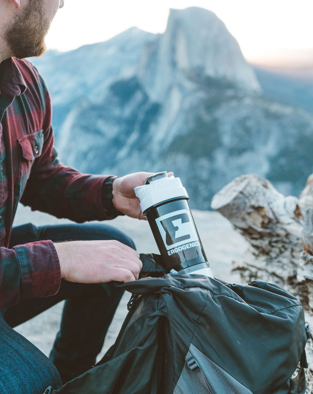 A man wearing a red plaid flannel shirt and blue jeans sits outdoors in a mountain setting, holding a black and white shaker bottle with the brand name 'PROGENSIS' next to a black backpack.