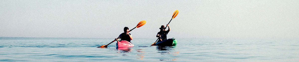 Two people kayaking on calm blue water with a pale sky, one in a pink kayak and the other in a green kayak, each holding an orange paddle.