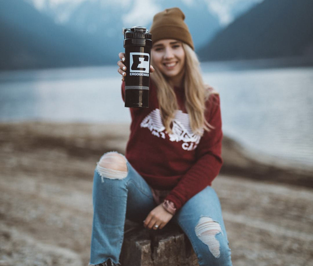 A woman sitting outdoors by a lake holds up a black water bottle with a white logo that reads 'Z Ergonomic'. She is wearing a brown beanie, a maroon sweatshirt, and ripped blue jeans, with mountains and water blurred in the background.