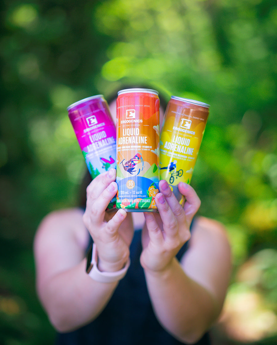 Three cans of Performance Nutrition liquid adrenaline drinks are held up by a person against a blurred green outdoor background. The cans feature vibrant, colorful labels for Green Tea, Coffee, and Orange Juice flavors.