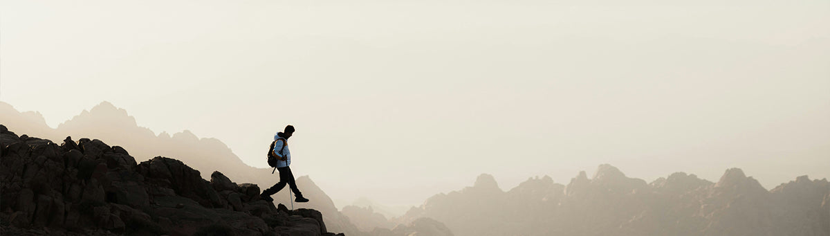 A hiker with a backpack walks along a rocky ridge against a misty mountain landscape under a pale sky.