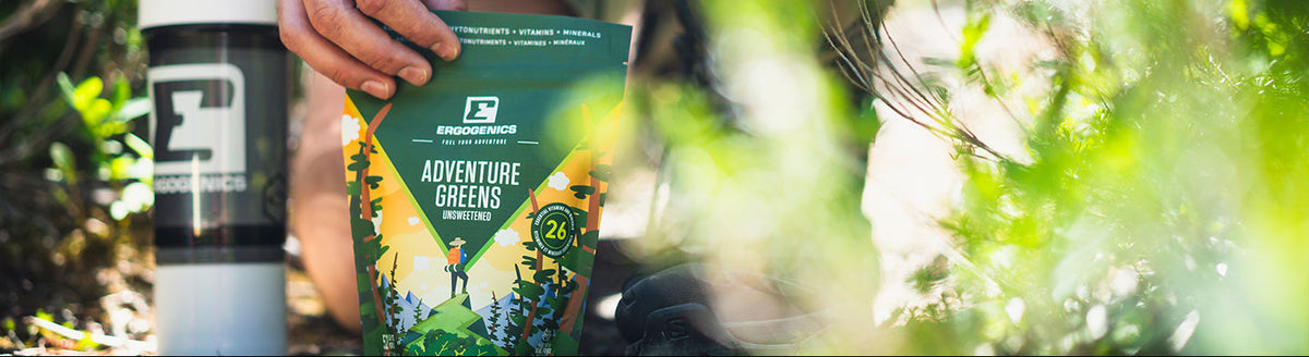 A person outdoors in a wooded setting holds a green ProQuinzai Adventure Greens snack pouch next to a white shaker bottle.