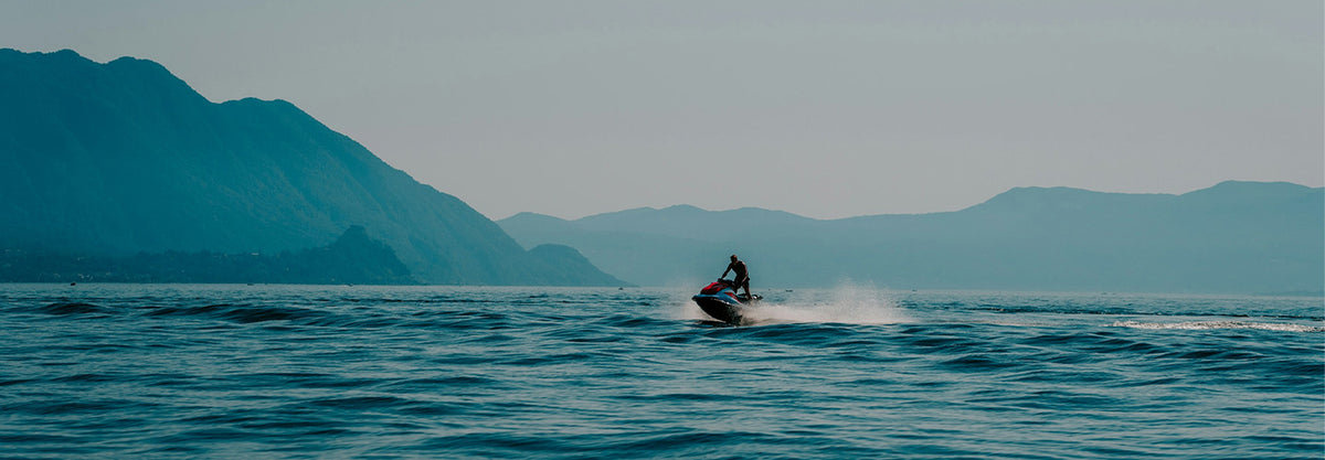 A person riding a jet ski across blue water with hazy mountains in the background under a pale sky.