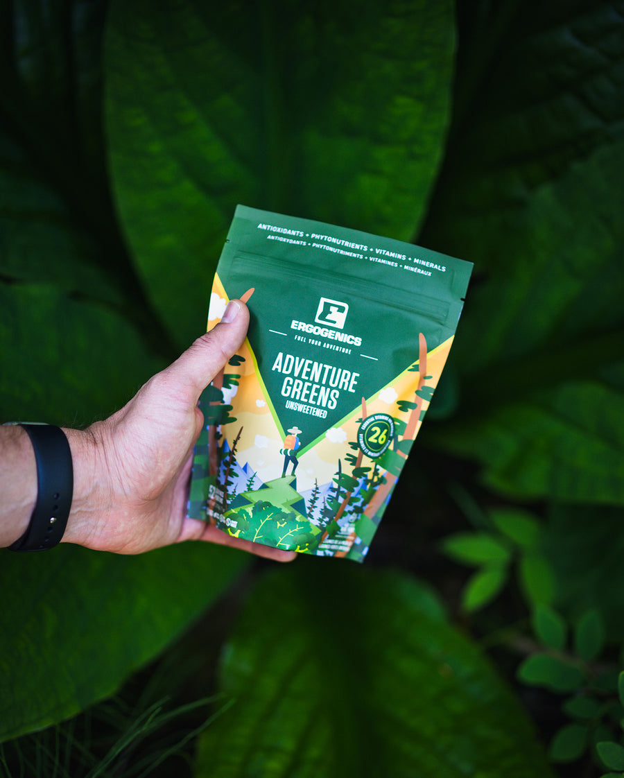 A hand holding a green bag of Ecoscience Adventure Greens Infused Flavors against a background of lush green leaves.