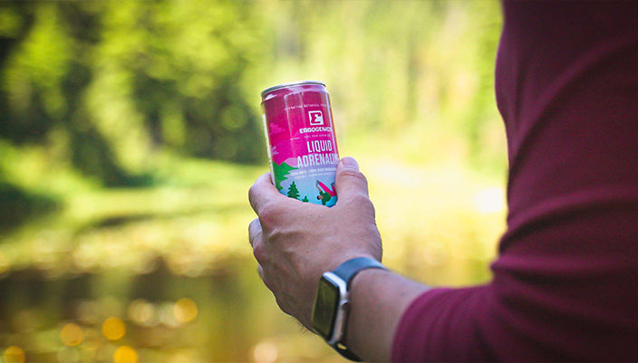 A person outdoors by a lake holds a can of BeringBottled Liquid Aggression in their hand. The background is a soft-focus green forest and water, and the person is wearing a maroon long-sleeve shirt and a smartwatch.