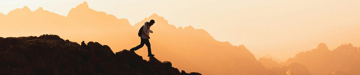 A silhouetted hiker with a backpack walks along a rugged mountain ridge against a warm orange and yellow misty mountain landscape at sunrise or sunset.
