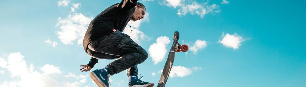 A skateboarder in a black hoodie and dark pants jumps against a bright blue sky with white clouds, with a skateboard visible below.