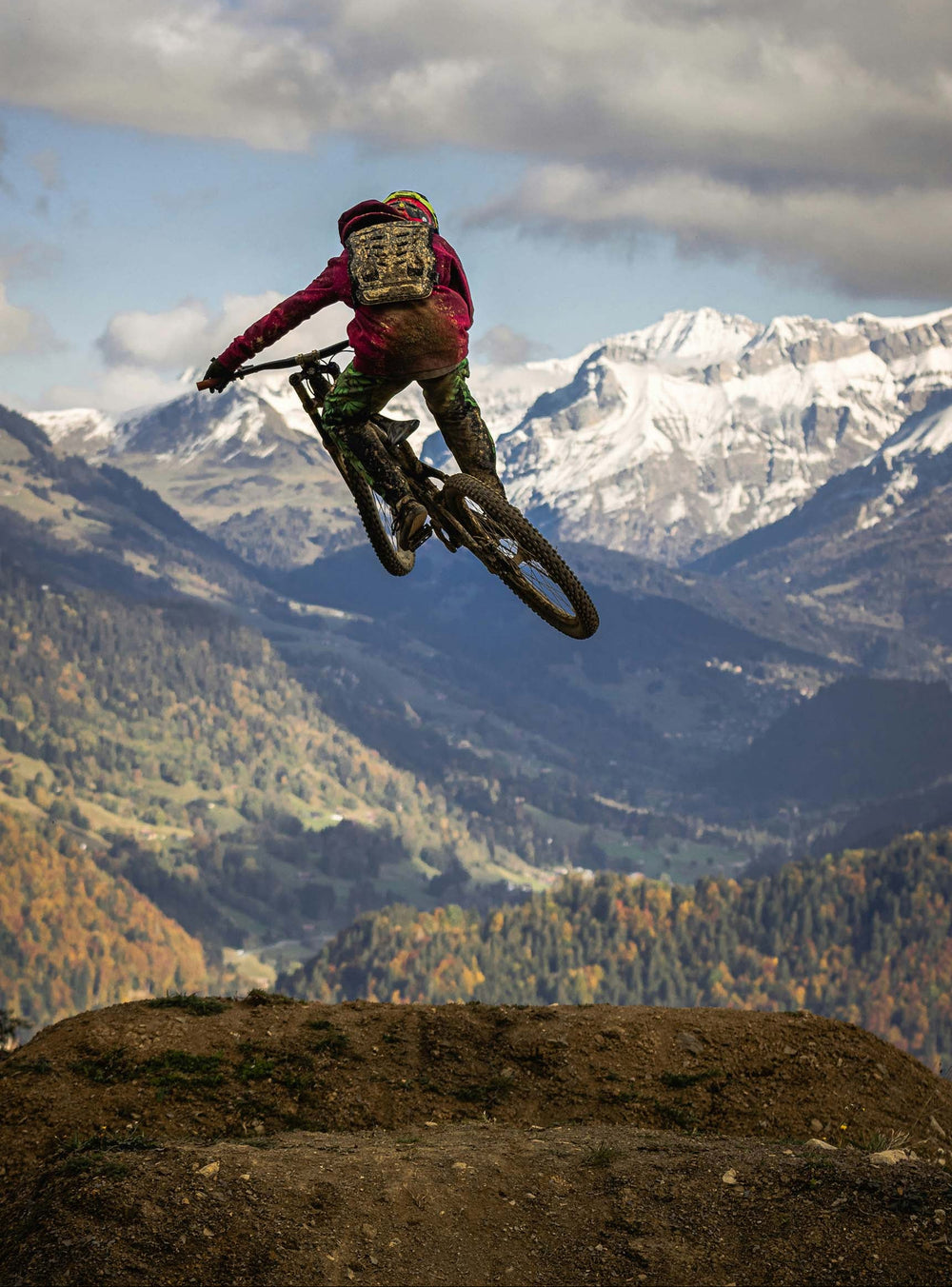 A mountain biker mid-air performing a jump over a dirt track, with snow-capped mountains and a cloudy sky in the background.