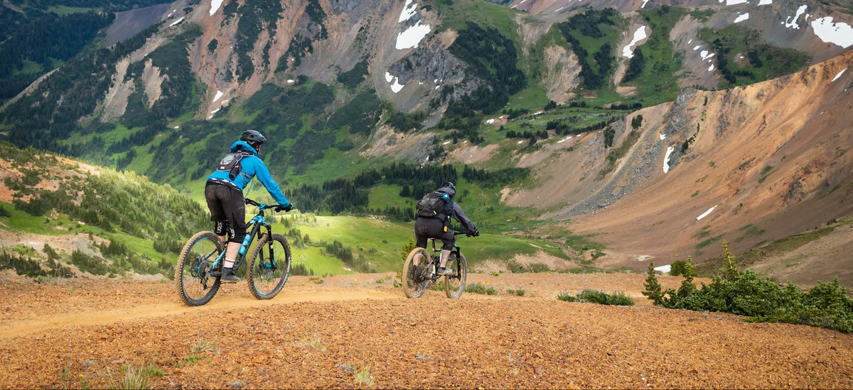 Two mountain bikers ride along a dirt trail in a vast mountain landscape with rocky peaks, green slopes, and distant snow-capped mountains.