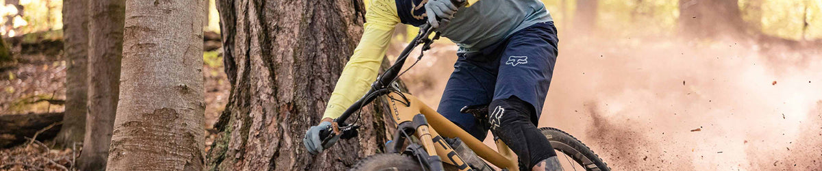 A mountain biker in a forest, wearing a yellow long-sleeve jersey, gray top, and navy shorts, riding past a large tree with dirt kicking up in the background.
