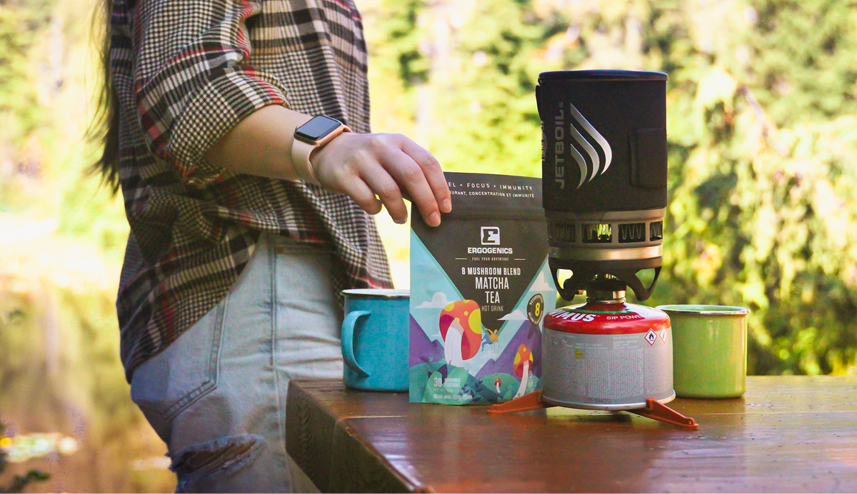 A person in a plaid shirt stands beside a wooden table outdoors, holding a box of Weddell's Mountain Blue Matcha Tea. On the table are camping mugs and a portable camping stove, with a blurred forest background.
