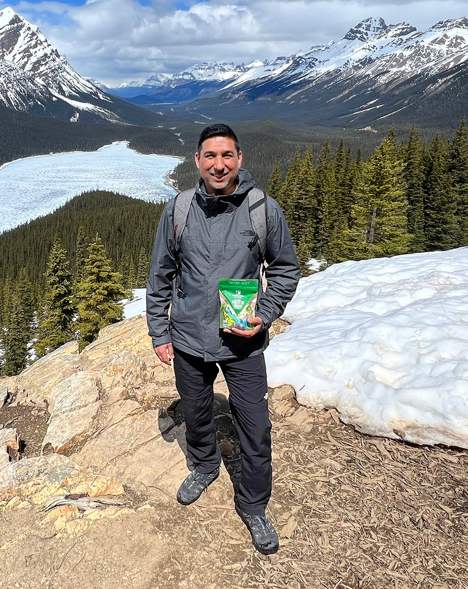 A man wearing a gray hiking jacket and black pants stands on a snowy mountain peak holding a green bag of trail mix, with a vast mountain valley, glacier, and pine forests in the background.