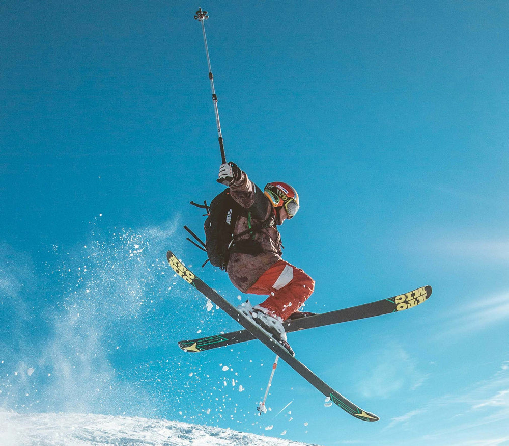 A skier mid-air performing a jump on a snowy mountain under a clear blue sky, wearing red pants, a dark jacket, and holding ski poles.