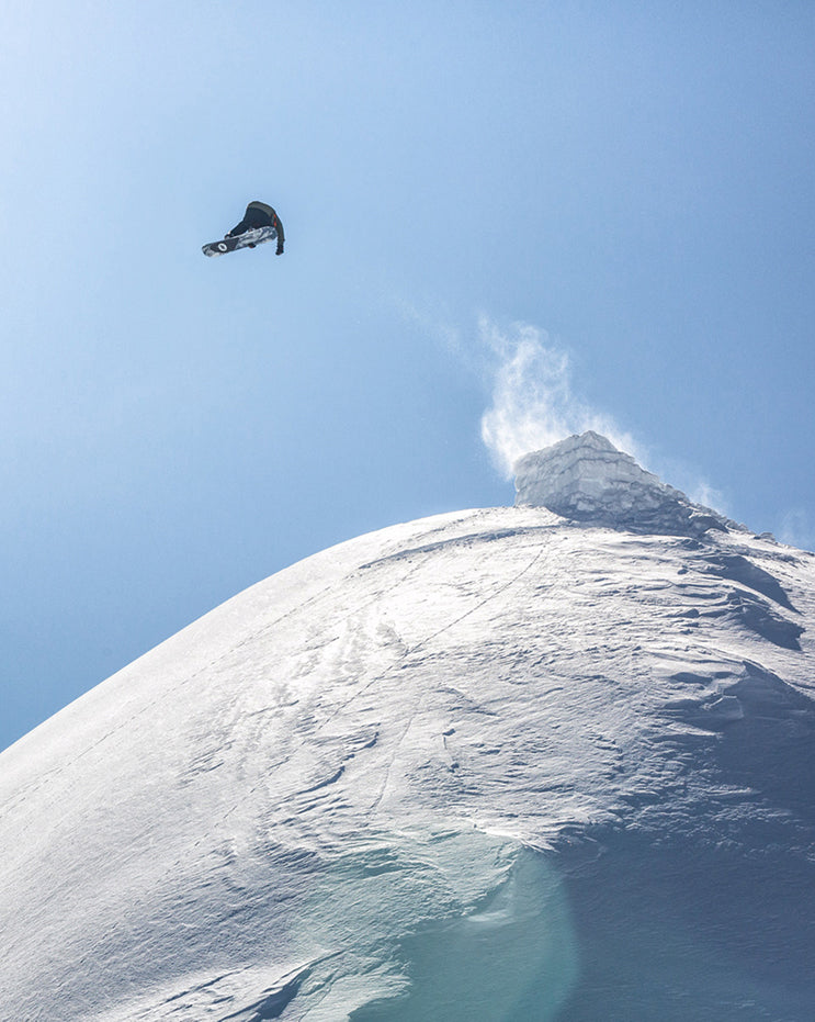 A snowboarder in a black and white outfit jumps high in the air against a clear blue sky, with a snow-covered mountain peak and a plume of snow in the background.
