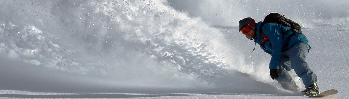 A snowboarder carving through deep powder snow, creating a large spray of white snow. The snowboarder is wearing a bright blue jacket, gray pants, and a black backpack.