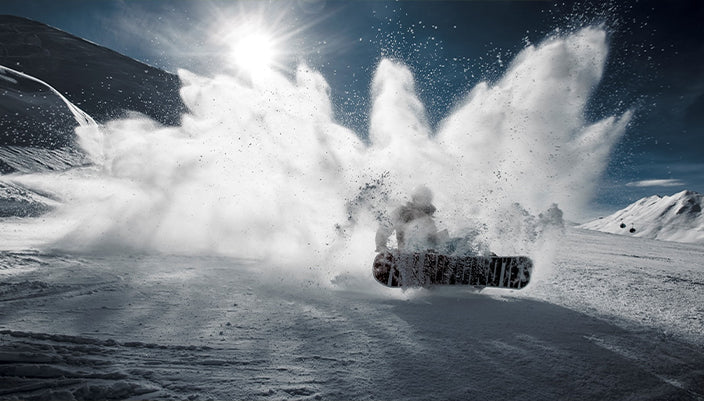 A snowboarder in motion creates a dramatic spray of snow in a snowy mountain landscape under a bright sun and blue sky.