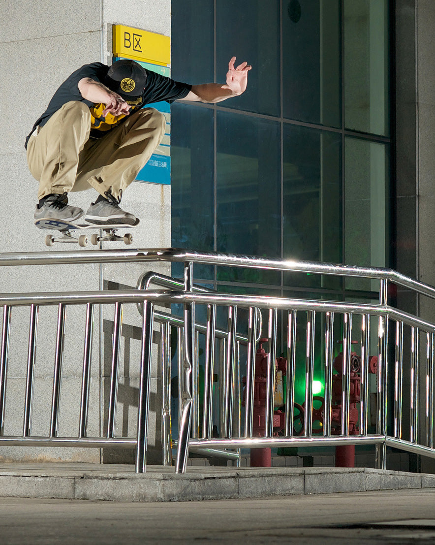 A skateboarder in a black t-shirt, beige pants, and a black cap performs a trick mid-air on a metal rail outside a modern glass building.
