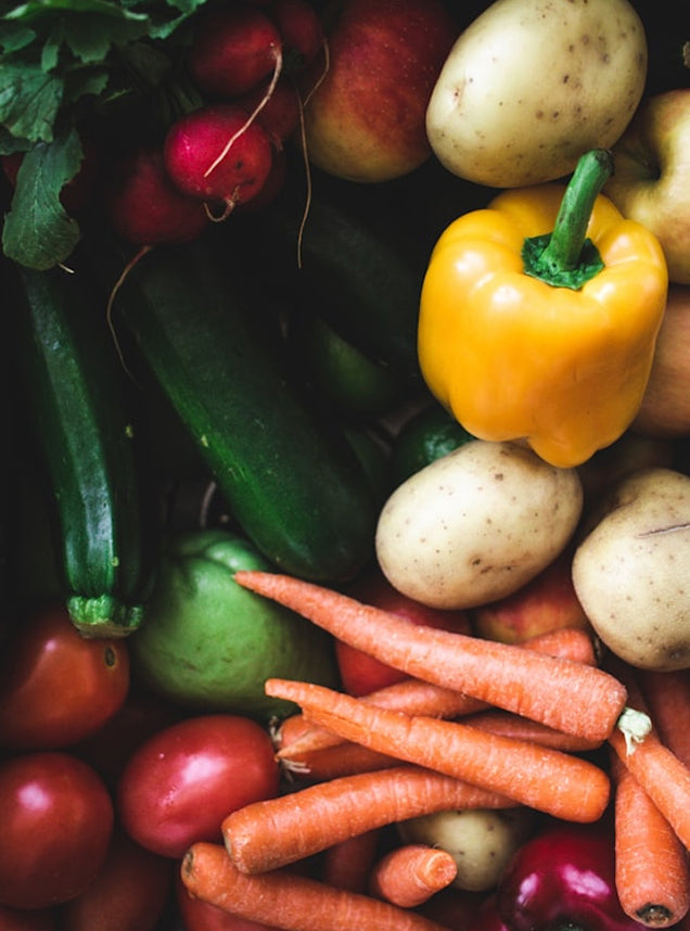 A close-up, top-down view of a colorful assortment of fresh vegetables, including orange carrots, a bright yellow bell pepper, green zucchinis, red radishes, and beige potatoes.