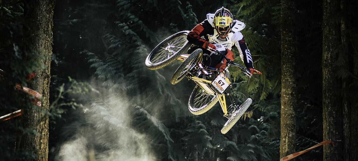A mountain biker in a forest jumps a dirt bike mid-air over a dusty track, wearing a white, red, and black racing suit with a yellow helmet.