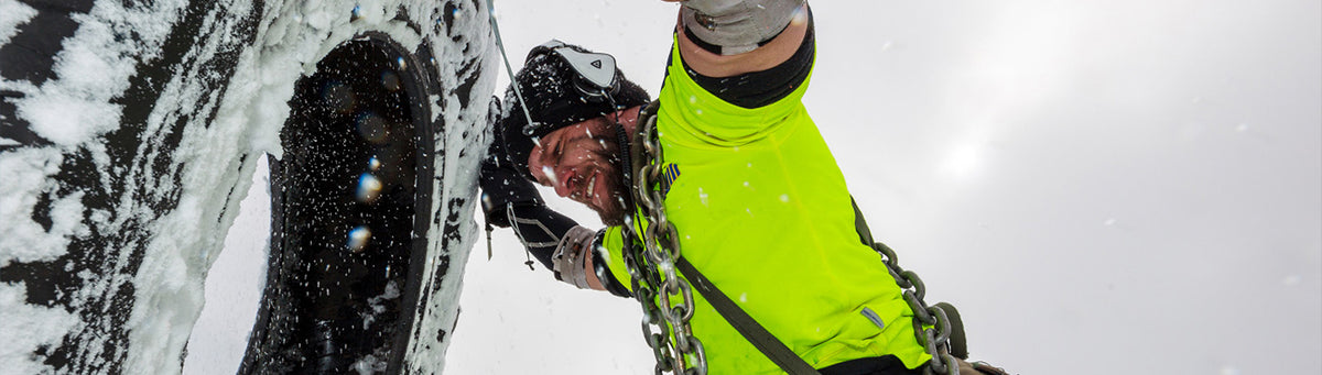 A person in a bright neon yellow high-visibility jacket and dark clothing is shown in a snowy outdoor setting, holding a heavy metal chain near a large dark object covered in snow.