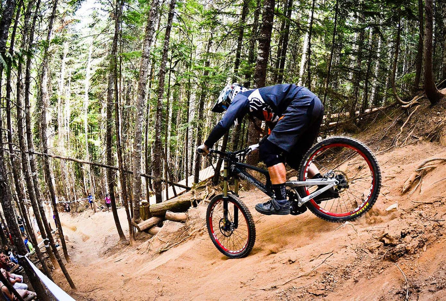 A mountain biker in a forest setting, leaning forward on a black full-suspension bike with red-rimmed wheels as it navigates a dirt trail.
