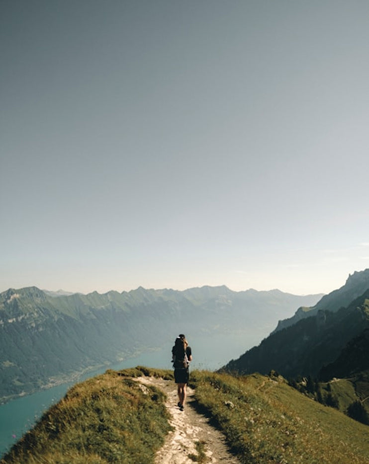 A person hiking on a narrow path along a mountain ridge overlooking a deep valley and lake, with layered green mountains fading into the distance under a pale sky.