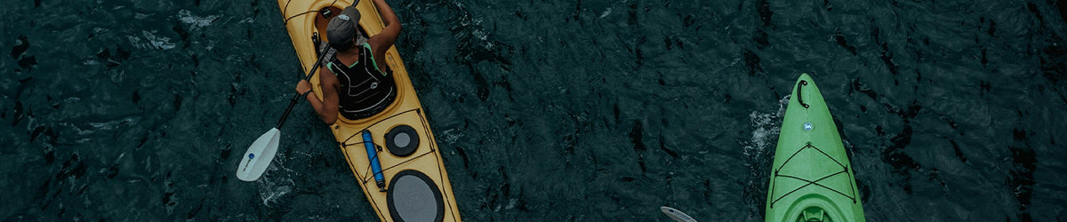 An aerial view of a person kayaking on dark blue water, with a yellow kayak containing the swimmer and another green kayak partially visible at the right edge.