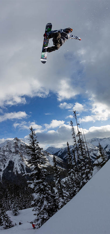 A snowboarder mid-air performing a jump in a snowy mountain setting with pine trees and a blue sky with clouds.