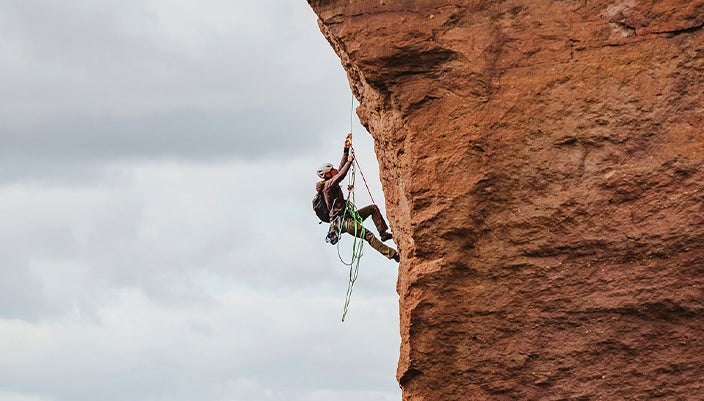 A rock climber suspended on a climbing rope against a large reddish-brown rock face, with an overcast cloudy sky in the background.