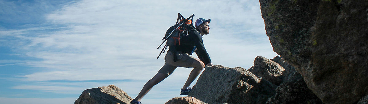 A trail runner wearing a black and blue backpack climbs rocky terrain under a blue sky with light clouds.