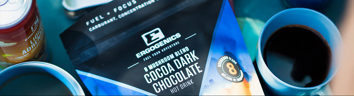 A bag of Ergogenics Cocoa Dark Chocolate hot drink mix on a teal tray, surrounded by coffee cups and beverage cans in a styled setting.
