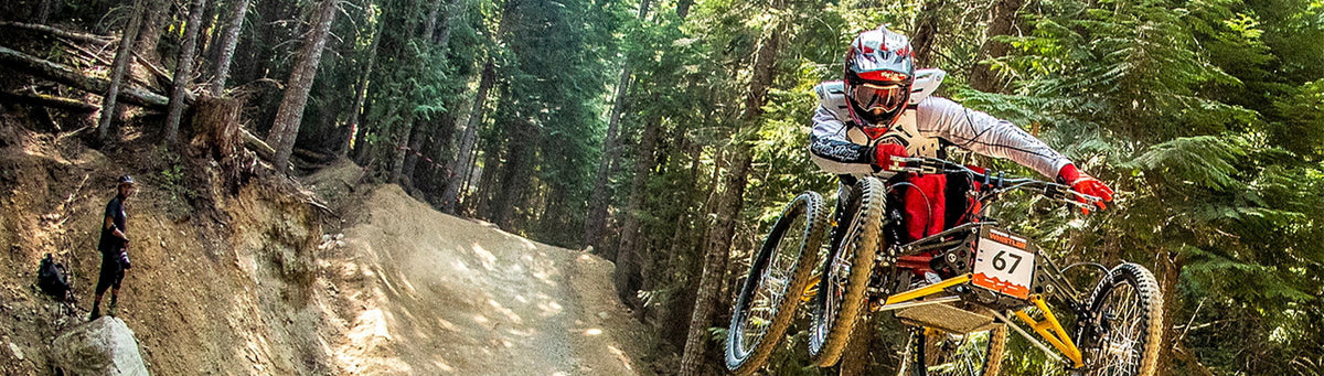 A mountain biker wearing a white helmet and red gloves rides a yellow and black bike down a dirt trail in a dense green forest. A second person is visible on the left side of the trail.
