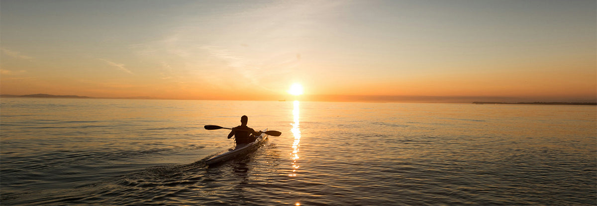 A silhouetted person paddling a kayak on calm water at sunset, with the sun low on the horizon and golden light reflecting across the surface.