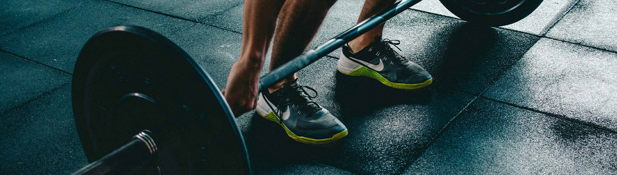 A close-up action shot of a person lifting a barbell in a gym, showing their legs, hands, and athletic shoes on dark rubber flooring.